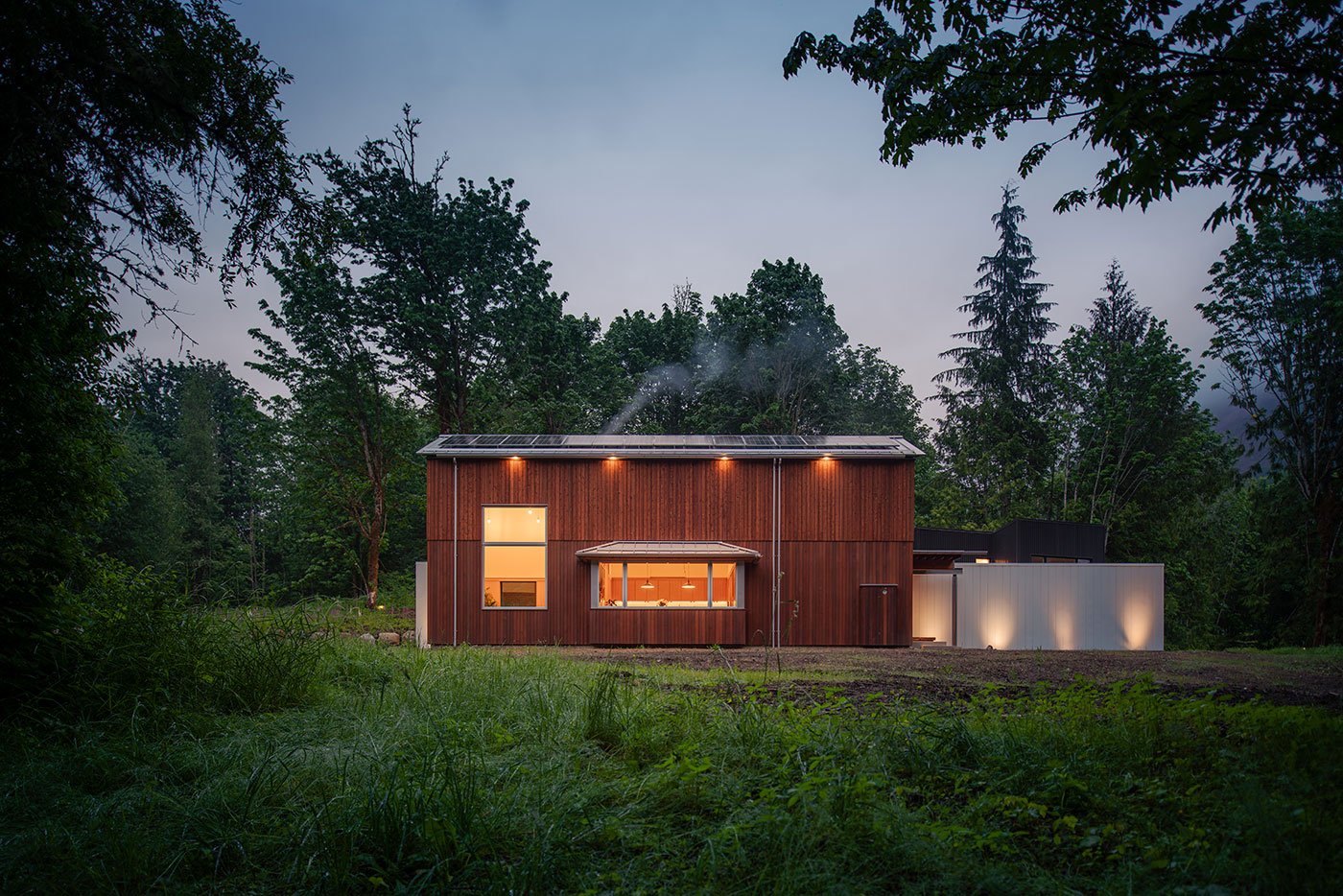 A barn-shaped home clad in vertical cedar with large-scale windows and a solar array on the roof is surrounded by meadow and forest.