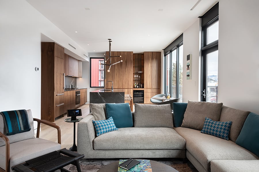 An interior shot of an open concept living room-kitchen with floor-to-ceiling windows and lots of wood cabinetry.