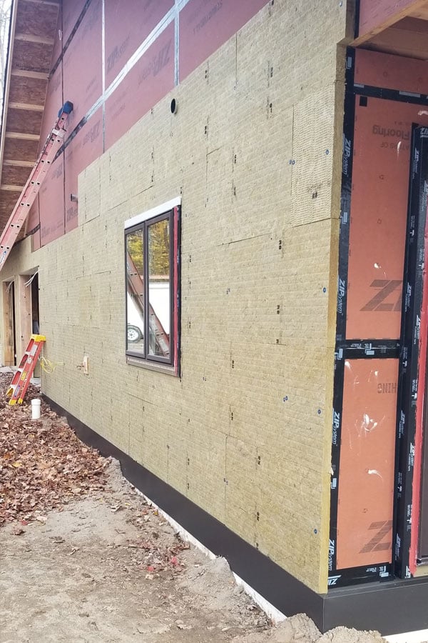 The exterior of a home under construction. The wall is partially covered with insulation panels and the lower section in a light yellow color and the upper section in red. There is also a window framed in view