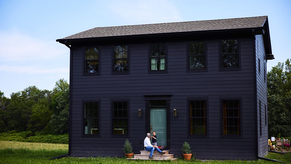 A man and young boy sit on the front steps of a black home with a green front door set in the middle of a grassy field with woods behind.