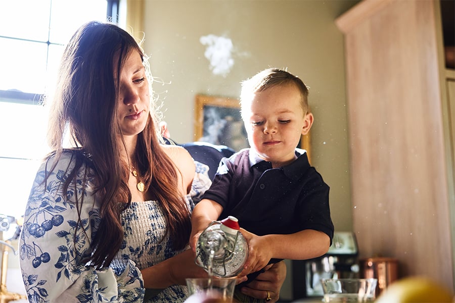 Inside a kitchen, a woman holds a young boy who is pouring water into a glass.