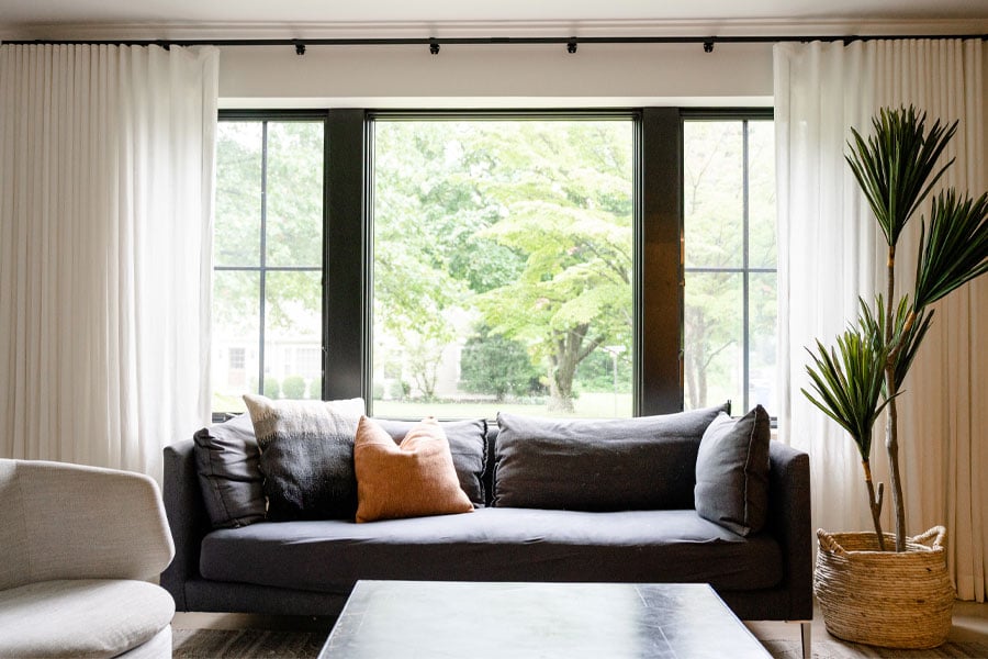 A living room couch sits in front of a picture window flanked by two casement windows with grilles and hung with white curtains.
