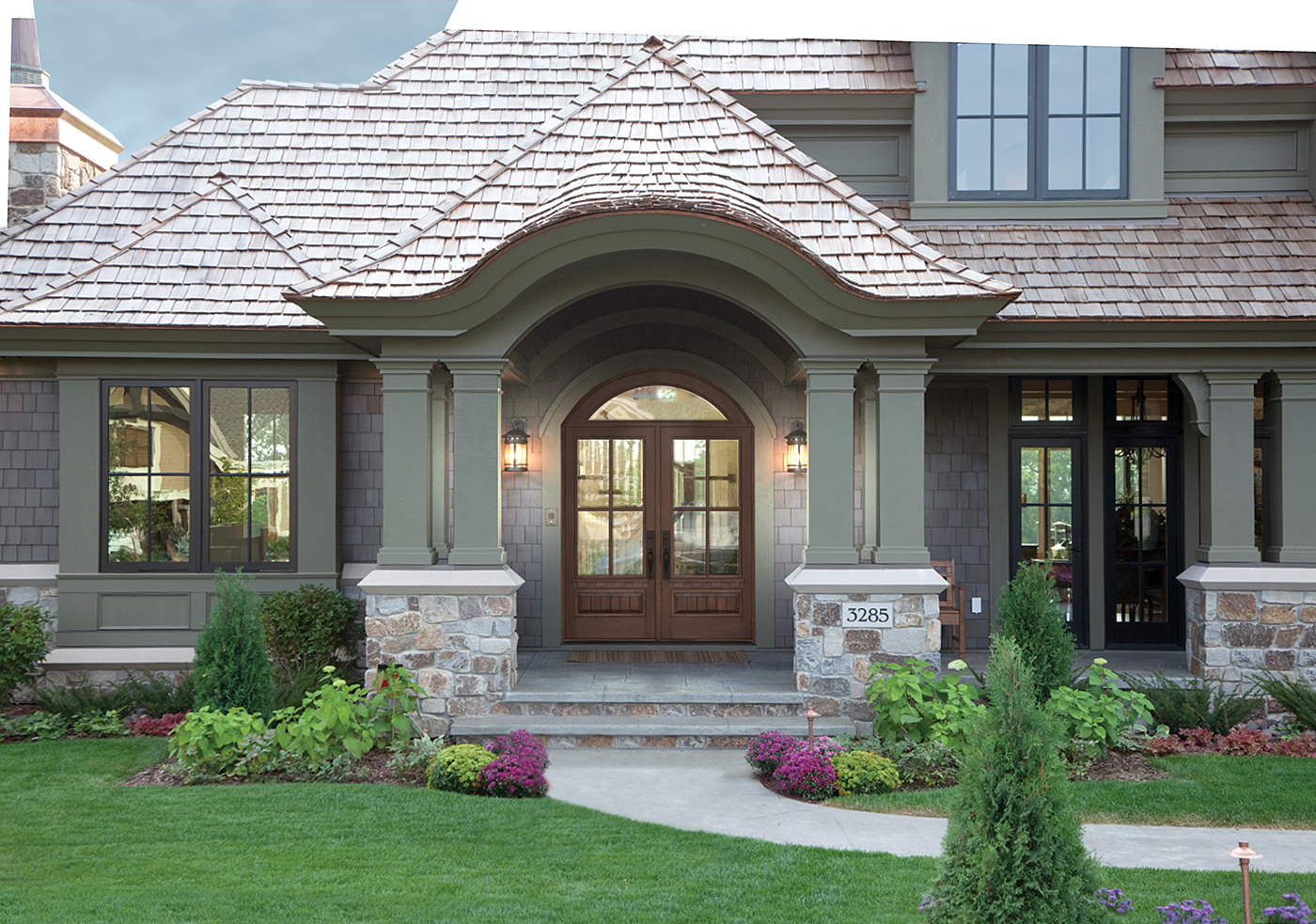 An entryway with a dark wood double front door featuring rectangle glass panels with an arched transom window above, lantern-style lights on either side, and wood shingles roof.