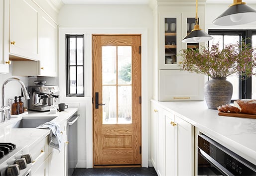 A kitchen with white painted cabinetry, white quartz counters, and a natural wood front door featuring a large glass panel with grilles.