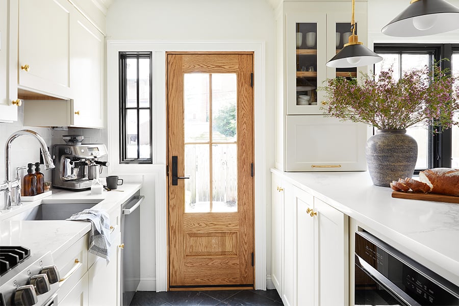 A kitchen with white painted cabinetry, white quartz counters, and a natural wood front door featuring a large glass panel with grilles.