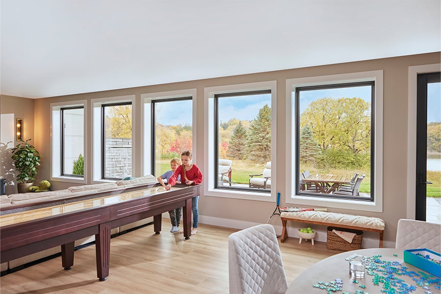 Two children play shuffleboard in a walkout basement with wood floors and a row of five picture windows behind them.