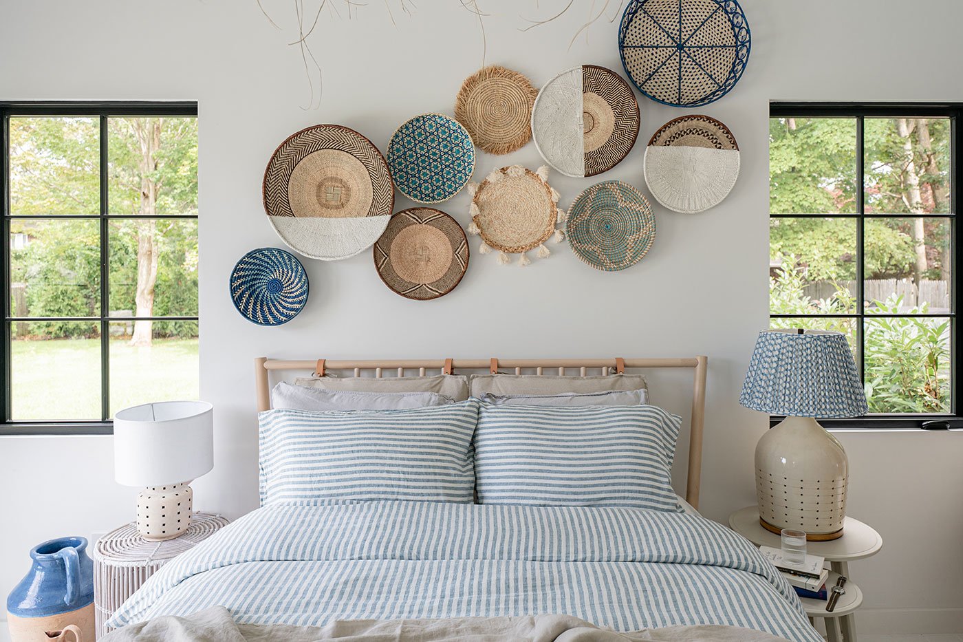 A bedroom with two black-framed windows with colonial grilles on either side of a bed dressed in blue-and-white striped linens with baskets hanging on the wall above.