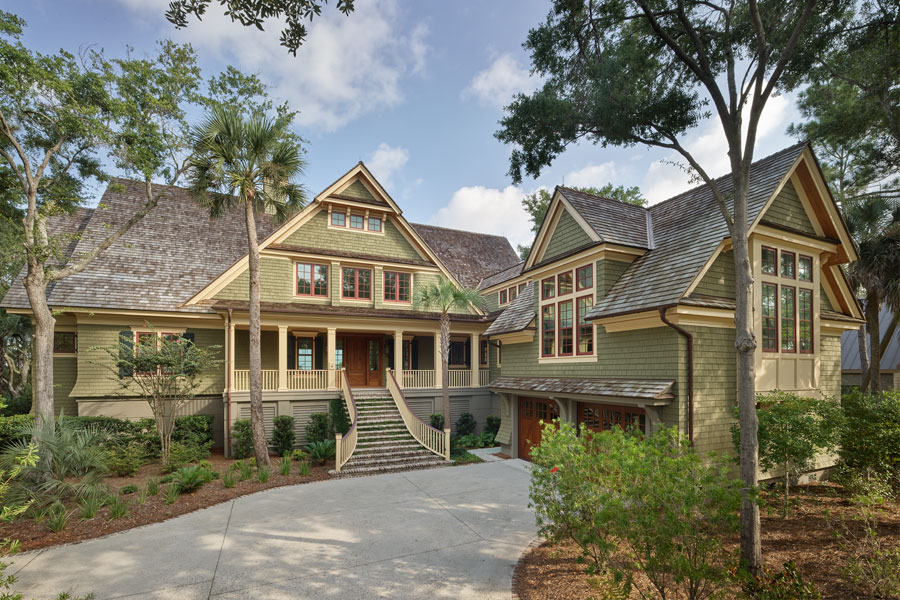 large, two-story Craftsman-style bungalow surrounded by lush greenery and tall trees. The house features a steeply pitched roof with shingles, multiple gables, and an inviting front porch with a wide staircase leading up to the entrance. The exterior is a mix of wood and stone, with numerous windows allowing natural light to pour into the home.  
