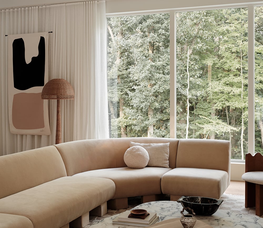 A living room with a checkered rug, beige couch and picture windows with white frames and curtains.