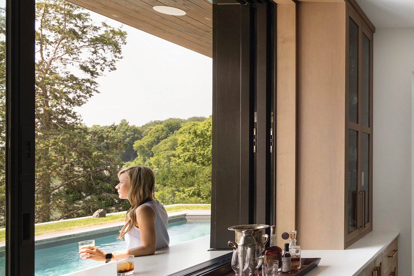 An open kitchen pass-through window sits flush with the countertop, which creates an outdoor bar where a woman sips her cocktail and looks out over the swimming pool.