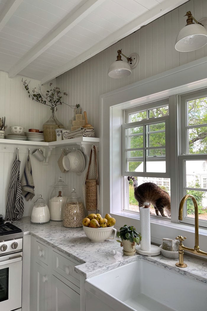 Two photos of a warm white cottage kitchen with wood-paneled walls, marble countertops, and a row of three double-hung windows above an apron sink.
