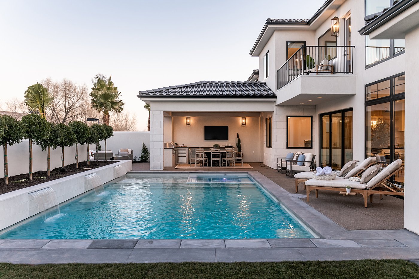 A Spanish-style home with black tile roof and black-framed windows and doors that’s oriented around the backyard swimming pool, outdoor kitchen, and lounge space.