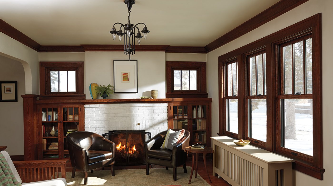 A craftsman living room with a fireplace, rich, dark-brown woodwork, and Andersen® 400 Series Woodwright Double-Hung Windows.